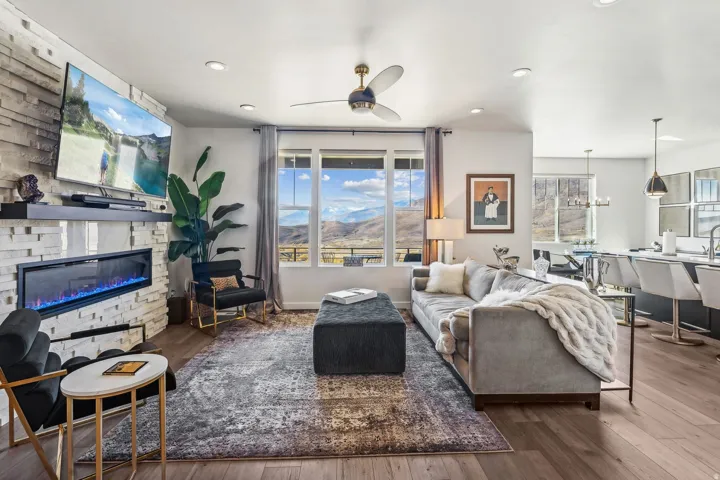 Spacious living area featuring a stacked stone fireplace with a built-in electric insert, wood-finish flooring, a ceiling fan, and large windows revealing a mountain landscape