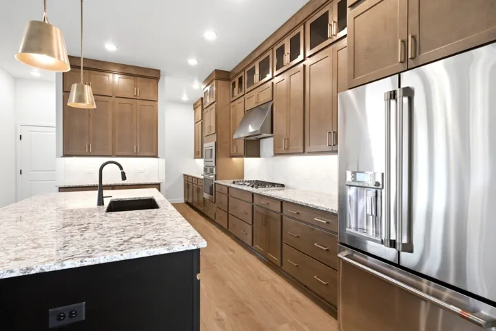 Gourmet kitchen featuring a spacious island with a black undermount sink and matte black faucet, light-colored stone countertops, wood-finish cabinetry, stainless steel appliances, and recessed lighting
