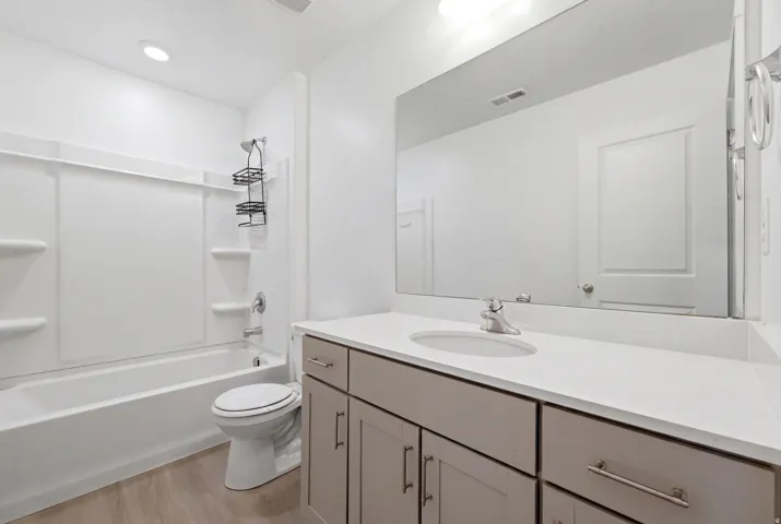 Bathroom featuring a white tub-shower combination with integrated shelving, a single-sink vanity with a light-toned countertop, and wood-finish flooring