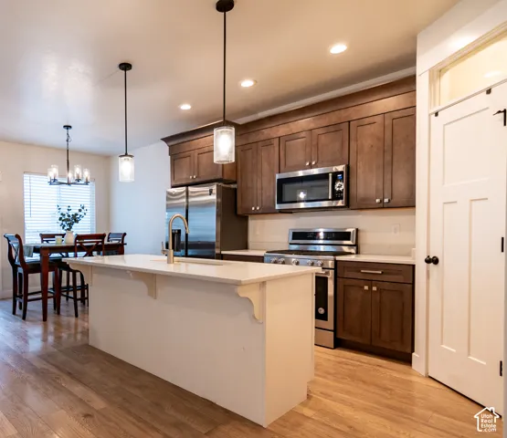 Open-concept kitchen featuring an island with a built-in sink, stainless steel appliances, wood-finish flooring, recessed lighting, and a white paneled door with a transom window