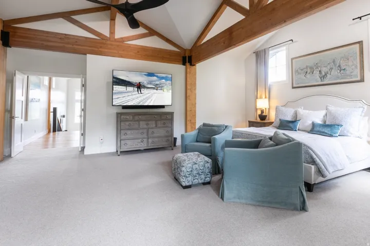 Vaulted ceiling with exposed wood beams, plush carpeting, white walls, and a window with a sheer curtain