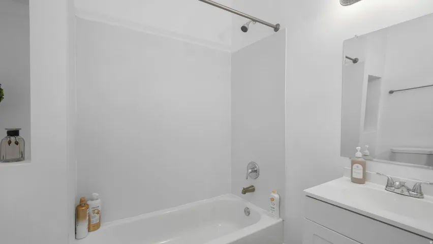 White bathroom featuring a bathtub with shower, chrome fixtures, built-in shelving, a vanity with an integrated sink, and a wall-mounted mirror