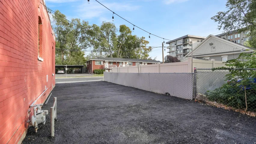 Paved outdoor area featuring a red brick building facade, string lighting, chain-link fence, and a beige privacy fence