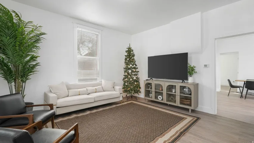 Bright living area featuring light wood-finish flooring, white walls, a single window with blinds, and an open doorway to another room