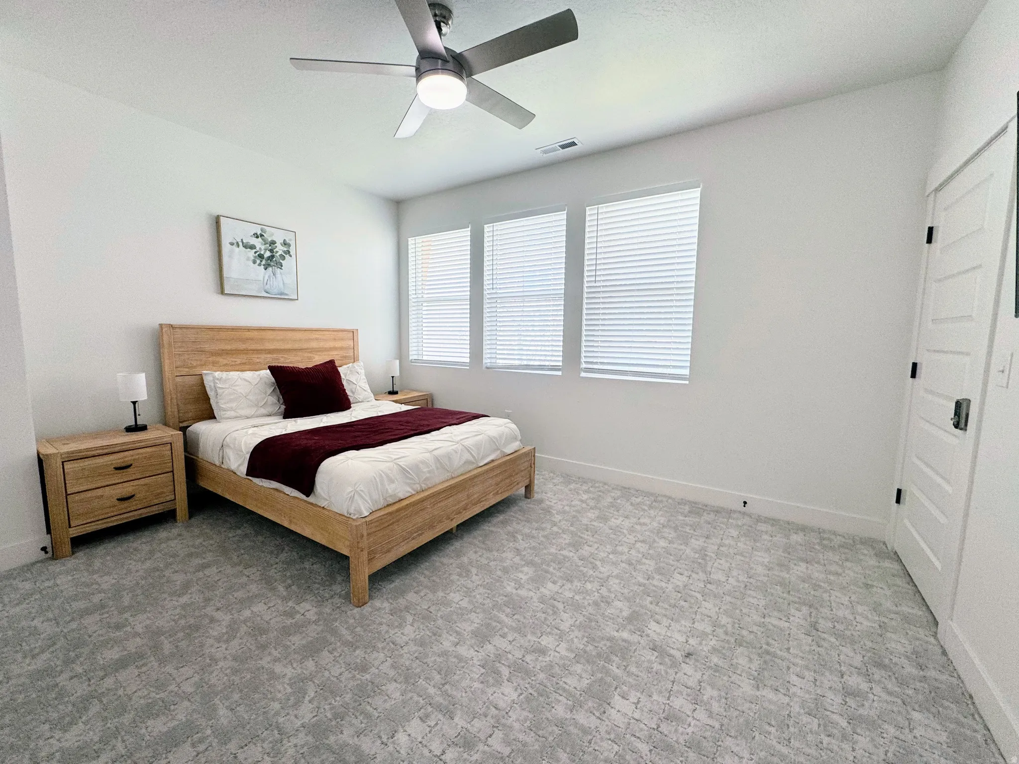 Bedroom featuring patterned carpeting, three windows with horizontal blinds, a ceiling fan with integrated lighting, and a paneled door with dark hardware