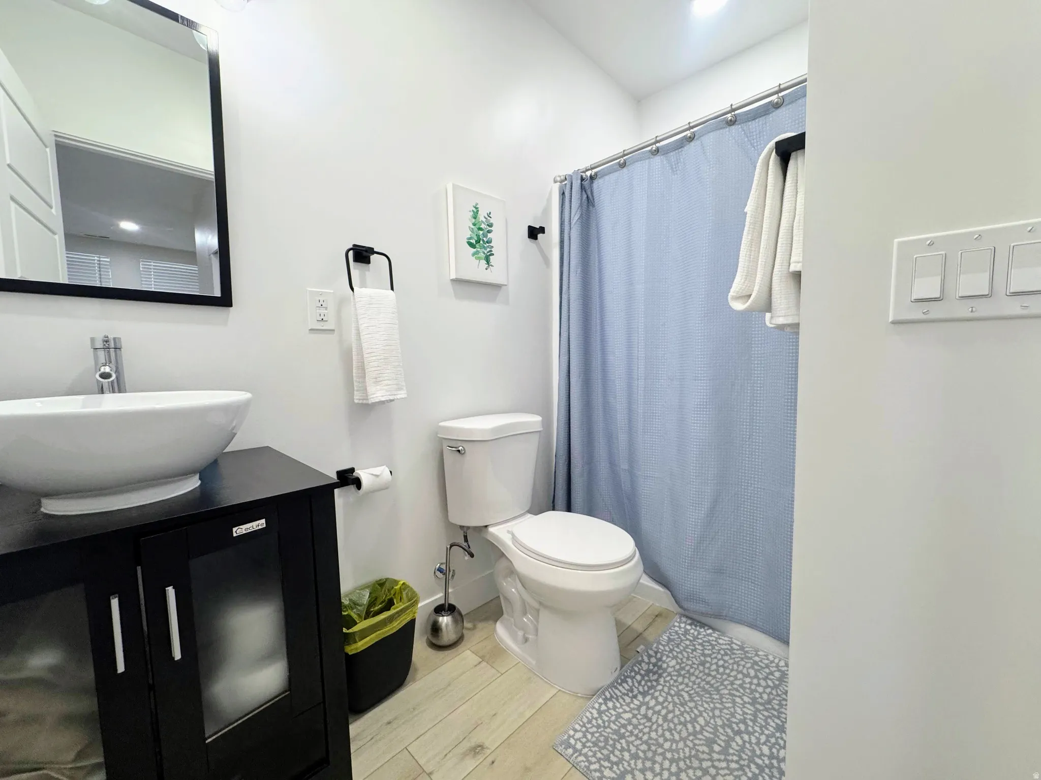 Bathroom featuring a black vanity with a vessel sink, a chrome faucet, a wall-mounted mirror, and wood-finish flooring