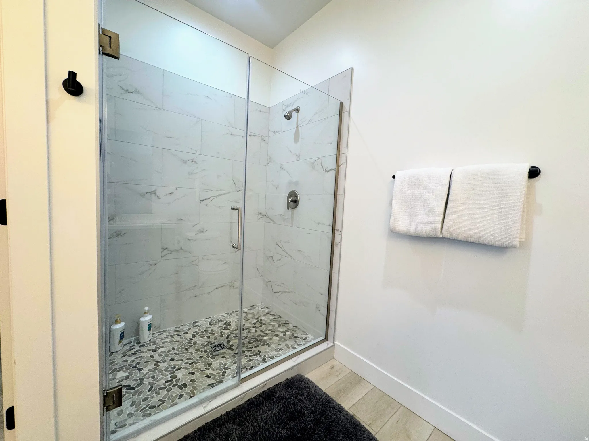 Glass-enclosed shower featuring large format wall tiling with subtle veining and a pebble-stone floor