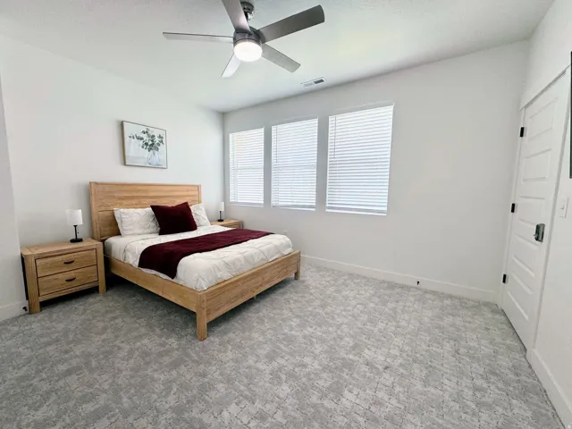 Bedroom featuring patterned carpeting, three windows with horizontal blinds, a ceiling fan with integrated lighting, and a paneled door with dark hardware