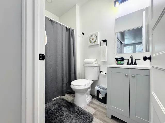 Bathroom featuring a single vanity with a gray cabinet base and white countertop, a wall-mounted mirror, a toilet, and wood-finish flooring