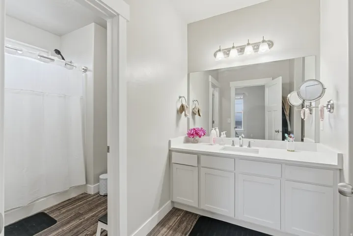 Bathroom featuring wood-finish flooring, a white vanity with multiple cabinets, a single integrated sink, a large wall-mounted mirror, and a shower with a chrome showerhead