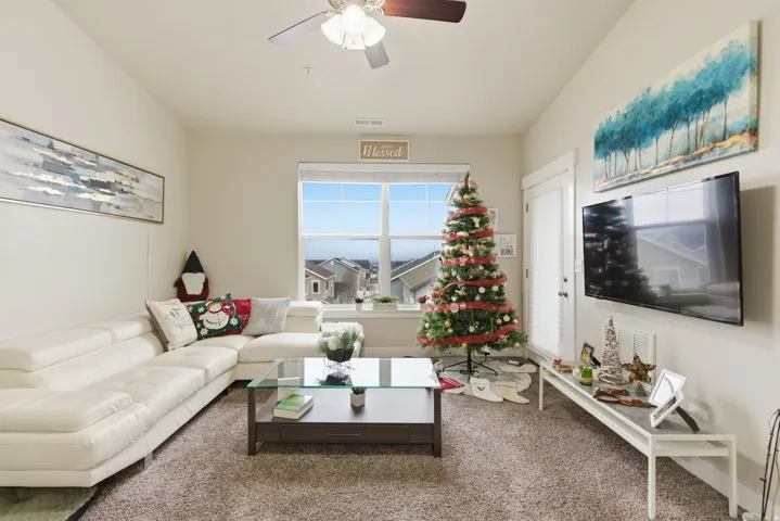 Living area featuring neutral-toned walls, textured carpeting, a window overlooking distant structures, and a ceiling fan with integrated lighting