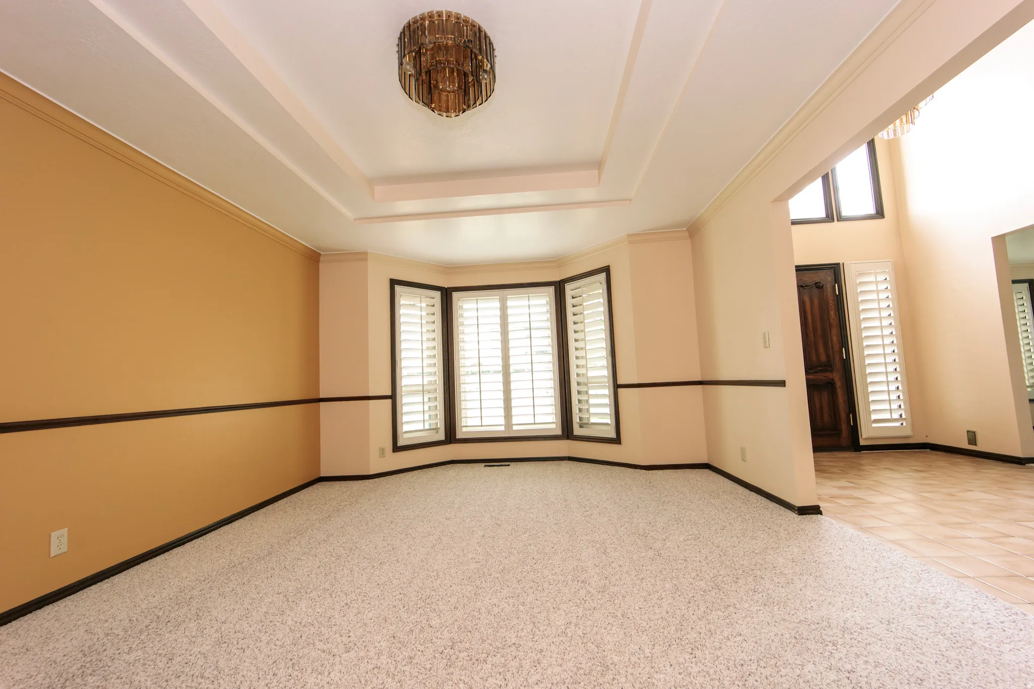 Recessed ceiling details with a central chandelier illuminate this room featuring a bay window with plantation shutters