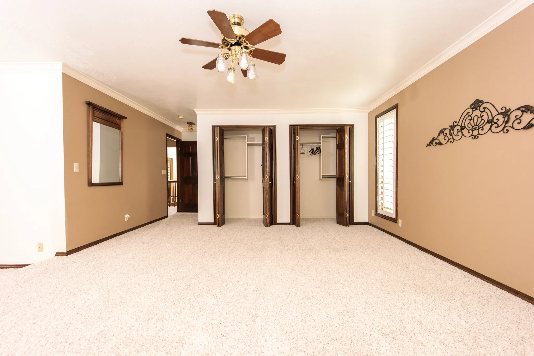 Spacious room featuring cream-colored carpeting, two double-door closets, crown molding, a ceiling fan with integrated lighting, and a window with white louvered shutters