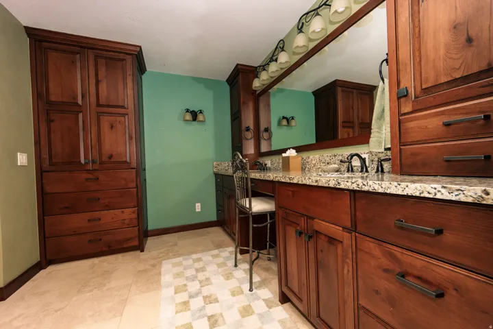 Bathroom vanity featuring dual sinks with dark bronze fixtures, granite countertop, expansive wall-mounted mirror with overhead lighting, and rich wood cabinetry