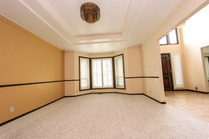 Recessed ceiling details with a central chandelier illuminate this room featuring a bay window with plantation shutters