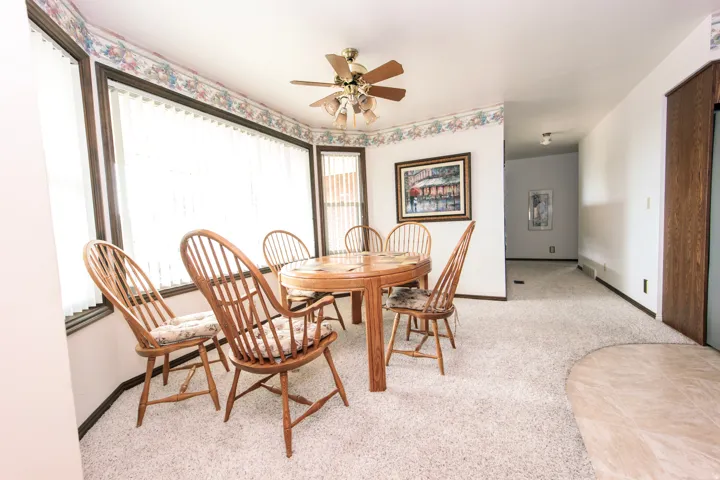 Bay window with vertical blinds, textured wall-to-wall carpeting, ceiling fan with integrated lighting, and a wood-finish built-in cabinet