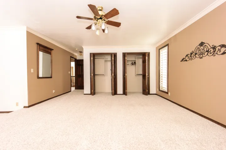 Spacious room featuring cream-colored carpeting, two double-door closets, crown molding, a ceiling fan with integrated lighting, and a window with white louvered shutters