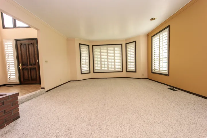 Spacious living area featuring light-colored carpeting, a bay window with plantation shutters, crown molding, and a brick-faced half-wall