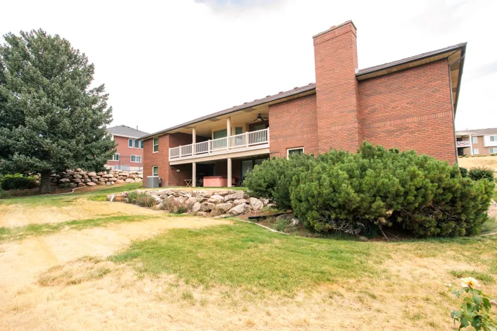 Brick exterior with a prominent chimney, upper-level balcony featuring white railings, lower-level patio area, and mature landscaping