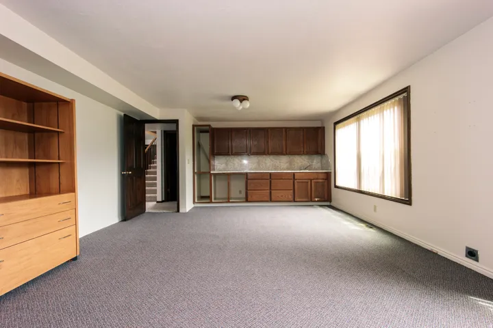 Expansive room featuring wood-finish built-in shelving, dark wood-finish cabinetry with light-toned backsplash, a wide window, and neutral carpeting