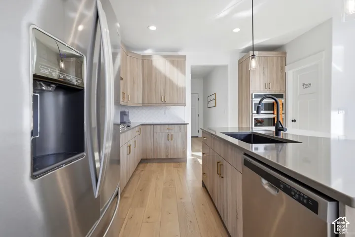 Stainless steel side-by-side refrigerator, wood-finish cabinetry, light wood-finish flooring, white subway tile backsplash, and a kitchen island with an integrated sink
