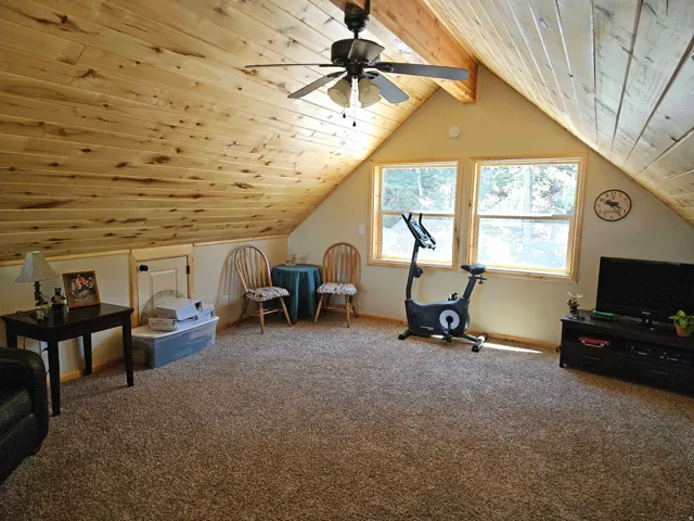 Carpeted loft space featuring an angled wood-paneled ceiling, exposed wood beams, and a ceiling fan