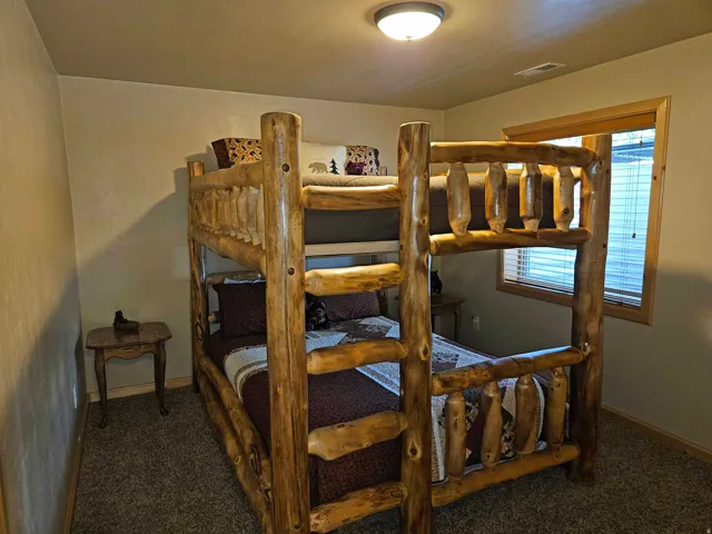 Bedroom featuring a natural wood bunk bed frame, neutral-toned carpeting, light beige wall paint, a window with horizontal blinds, and a flush-mount ceiling light