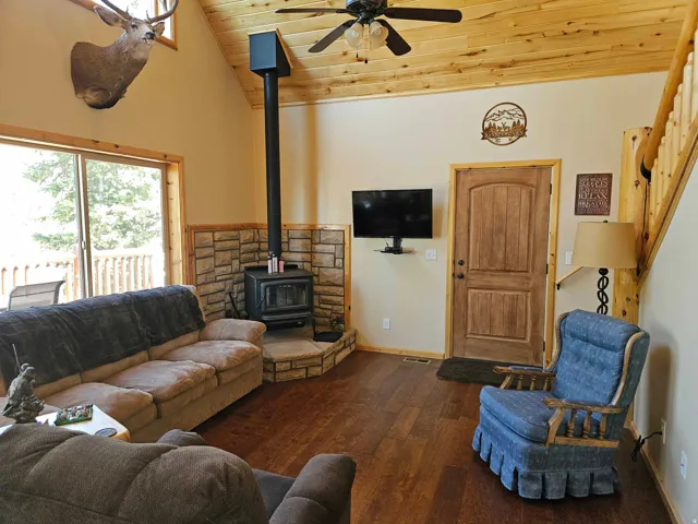 Living area featuring a stone-clad wood-burning stove with a black stovepipe, wood-finish flooring, and a vaulted ceiling with natural wood paneling