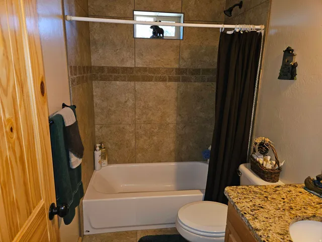 Bathroom featuring a tiled tub and shower surround, a window within the shower, a white bathtub, a toilet, and a vanity with a speckled countertop