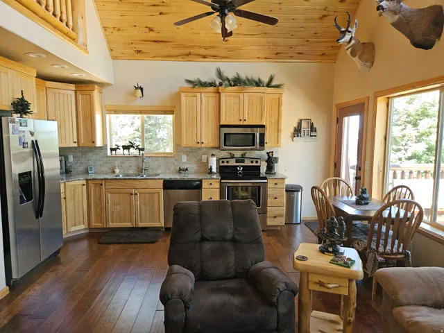 Open concept living space featuring wood-finish flooring, a vaulted wood-paneled ceiling, and a kitchen with light wood cabinetry, stainless steel appliances, and a tiled backsplash