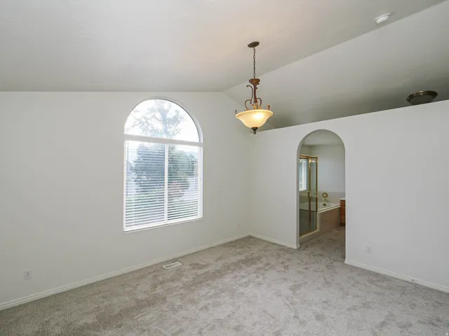Vaulted ceiling, arched window with blinds, carpeted flooring, and an arched doorway leading to a bathing area