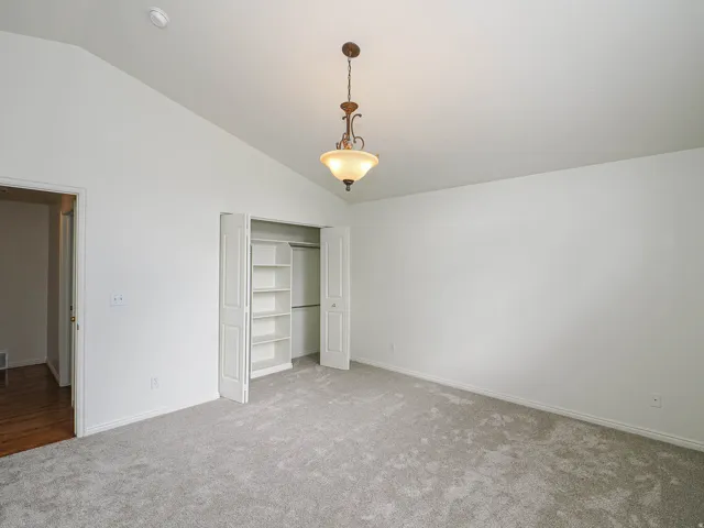 Vaulted ceiling with a central pendant light fixture, soft gray carpet, white walls, and a bi-fold closet with built-in shelving