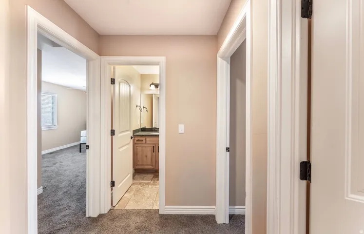 Hallway featuring beige walls, white trim, and gray carpet, with an arched doorway leading to a room with a window and a second doorway revealing a bathroom with a wood-finish vanity, dark countertop, and tiled flooring