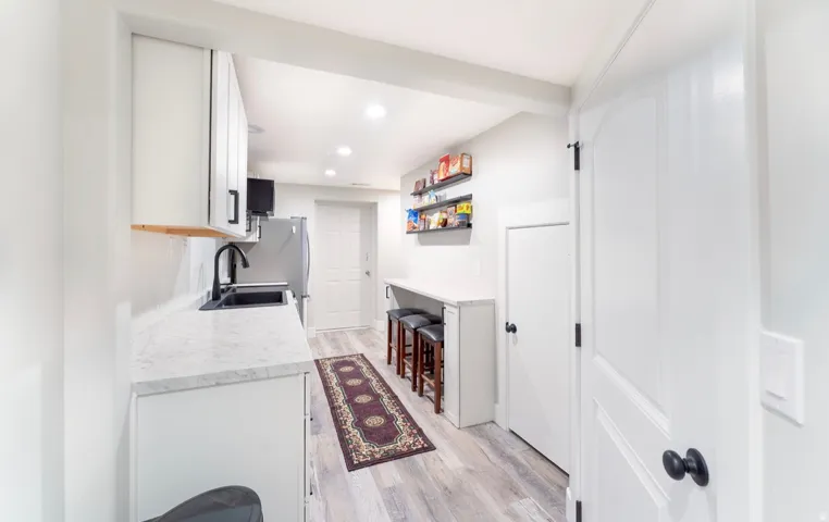 Compact kitchen area featuring light wood-finish flooring, white cabinetry with black hardware, a black sink with a matte black faucet, and light-toned countertops