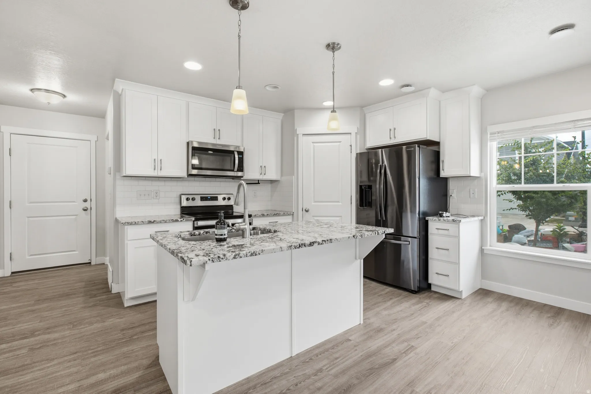 Contemporary kitchen featuring a central island with a sink, white cabinetry, stainless steel appliances, light-colored countertops, and wood-finish flooring