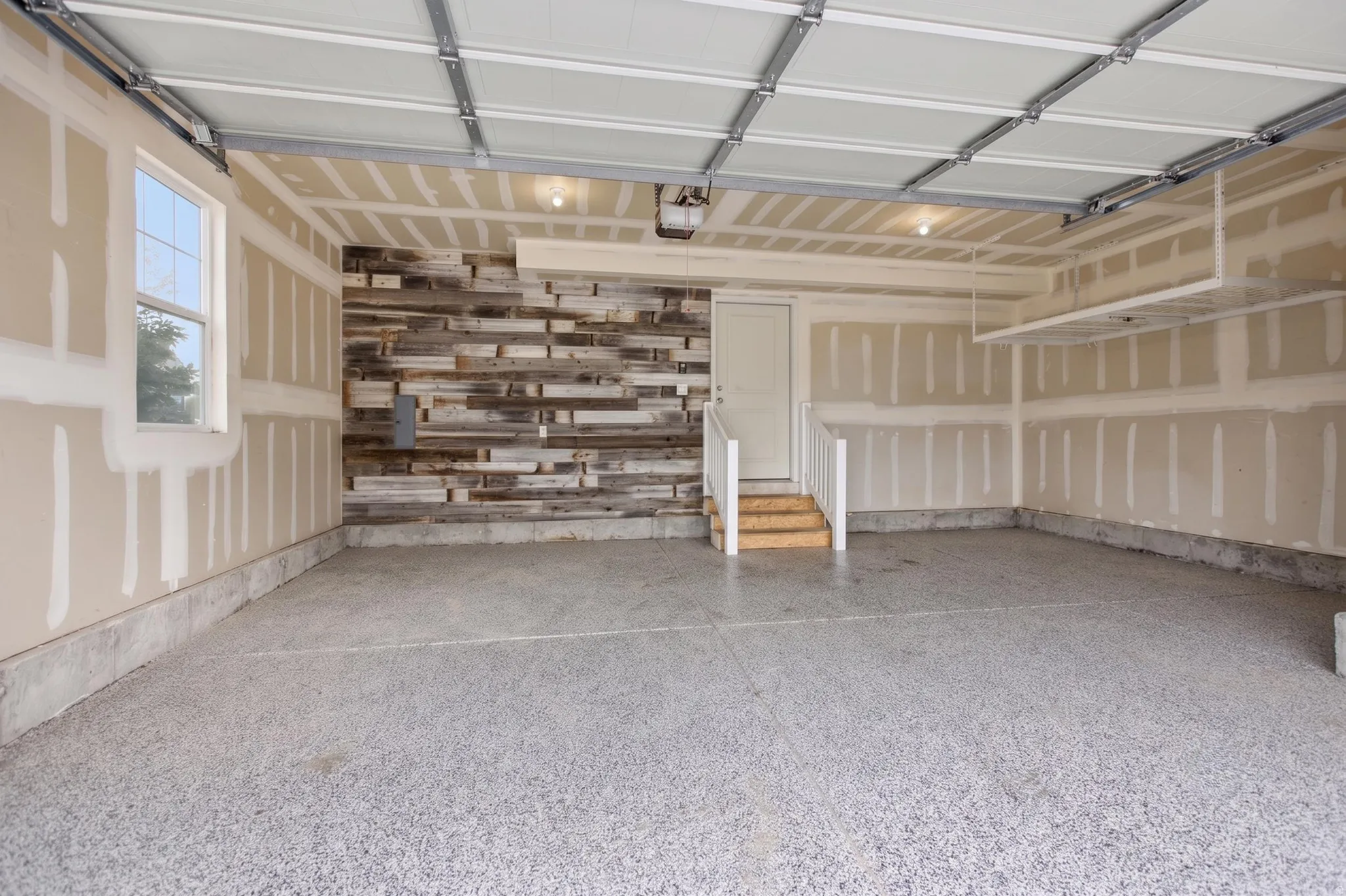 Spacious garage featuring a durable epoxy floor, a window providing natural light, an overhead storage rack, a built-in reclaimed wood accent wall, and interior access via a staircase with white railings