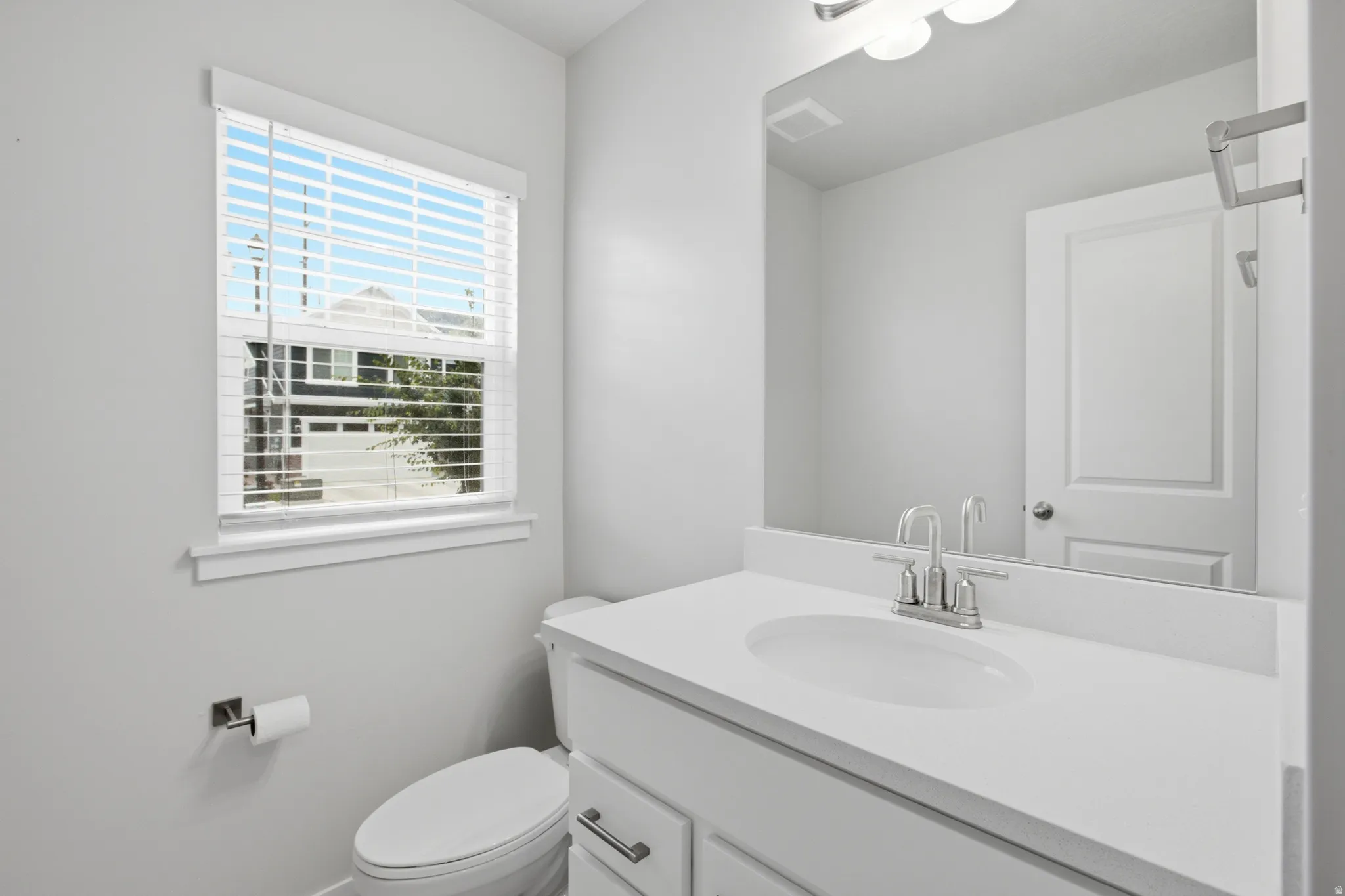 Bathroom featuring a single window with blinds, a vanity with a white countertop and undermount sink, a chrome faucet, a wall-mounted mirror, and a white toilet