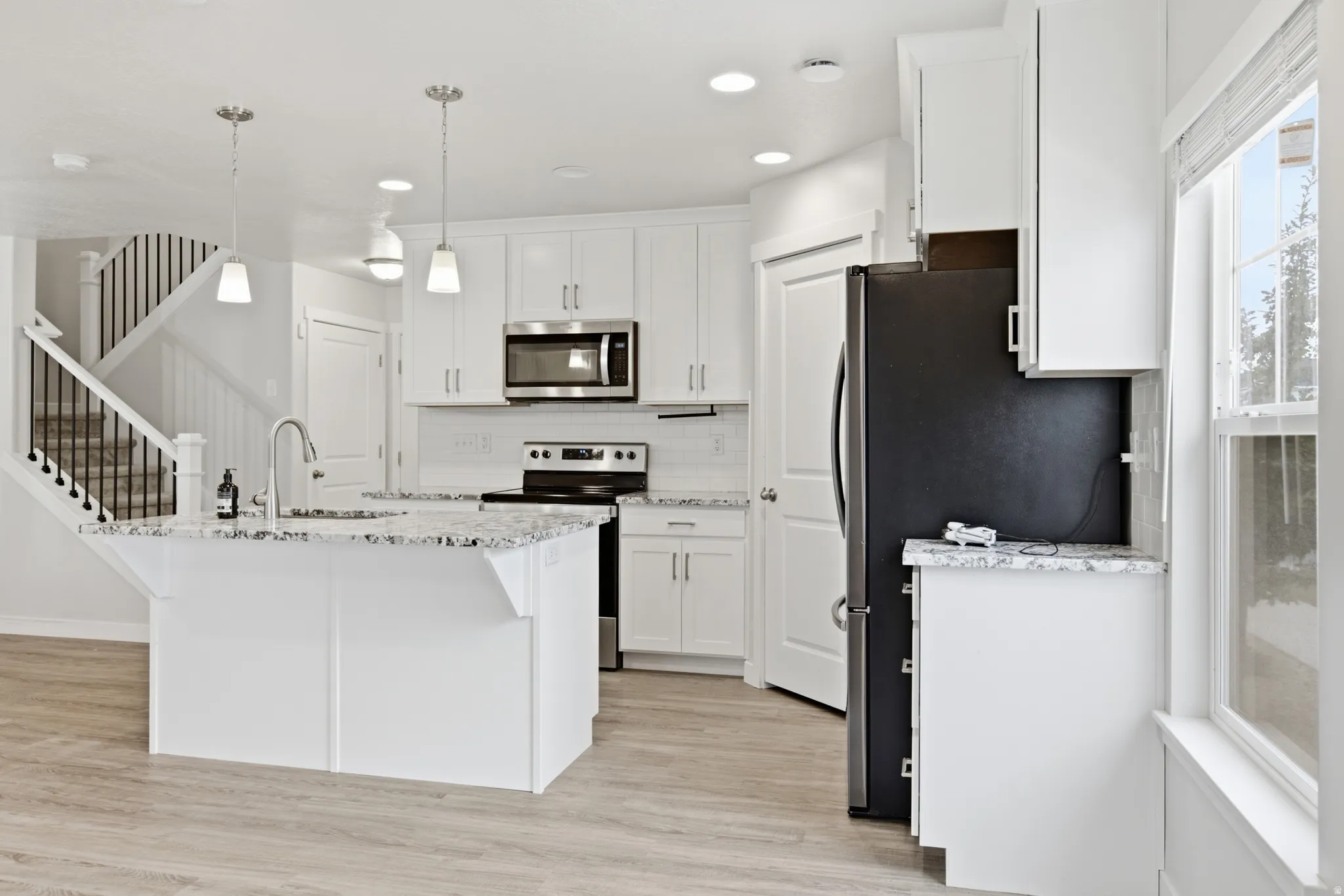 Contemporary kitchen featuring an island with a stainless steel sink and granite-style countertop, white shaker cabinetry, stainless steel appliances including a microwave and range, and wood-finish flooring