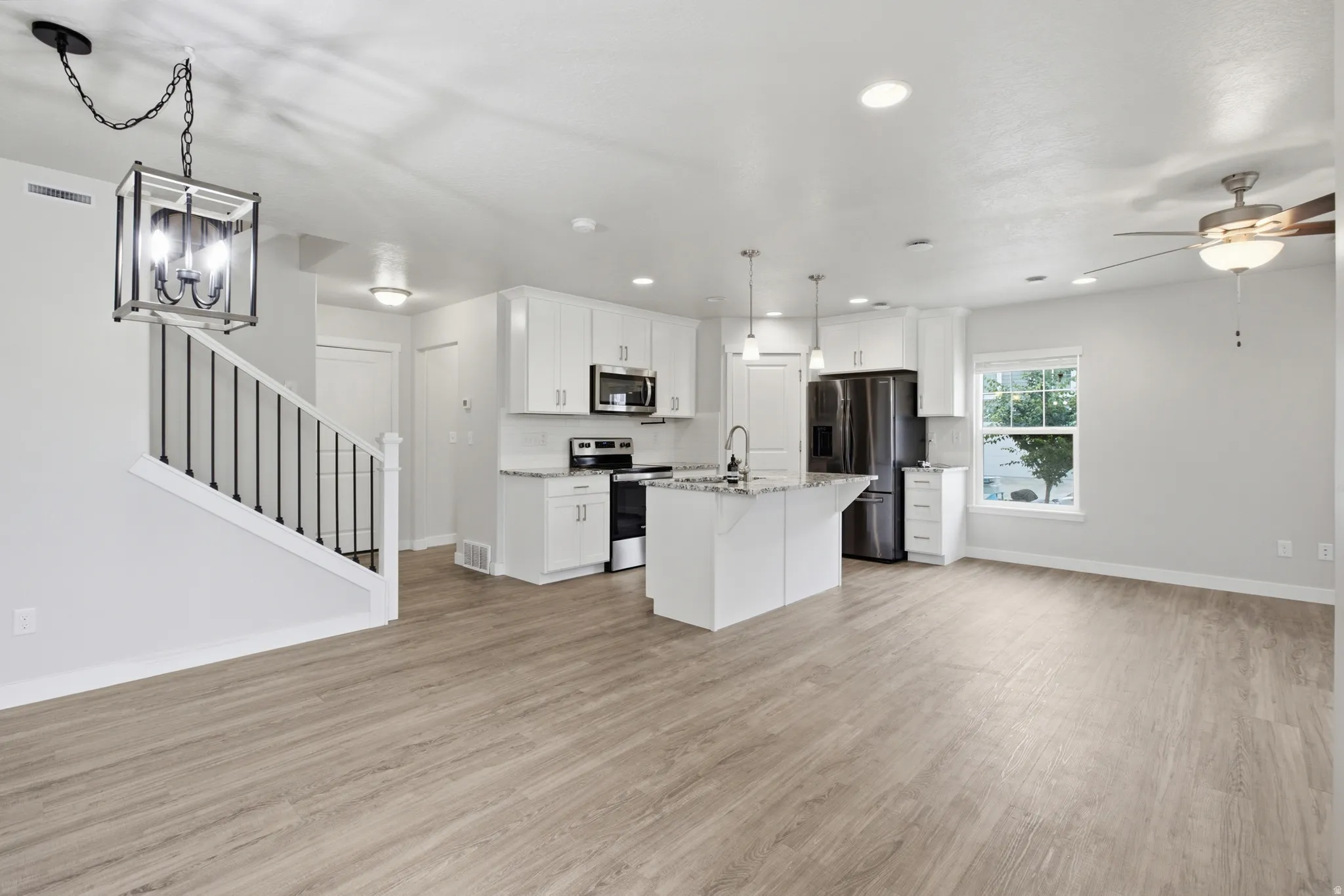 Open-concept living area featuring wood-finish flooring, a contemporary kitchen with white cabinetry and stainless steel appliances, a kitchen island with pendant lighting, a ceiling fan, and a staircase with an iron spindle railing and a decorative chand
