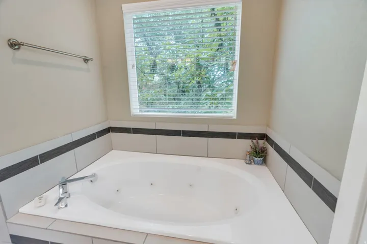 Corner jetted bathtub featuring a chrome faucet, tile surround with a dark accent band, and a window with blinds