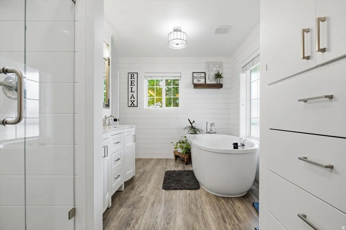 Spacious bathroom featuring a freestanding soaking tub, wood-finish flooring, a white vanity with a light-colored countertop, white shiplap walls, and a glass-enclosed shower with white subway tile