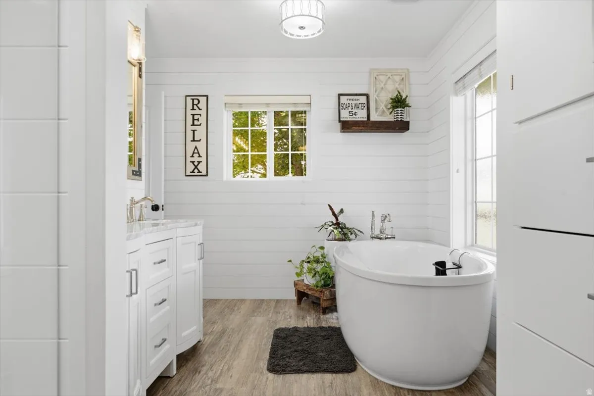 Bathroom featuring a freestanding oval tub, white shiplap walls, wood-finish flooring, a white vanity with a light-colored countertop, and a window with natural light