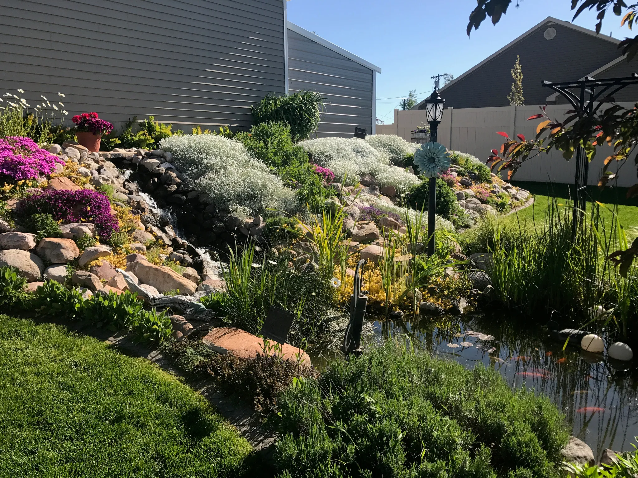 Landscaped yard featuring a multi-tiered rock garden with diverse plantings, a flowing waterfall, and a tranquil pond