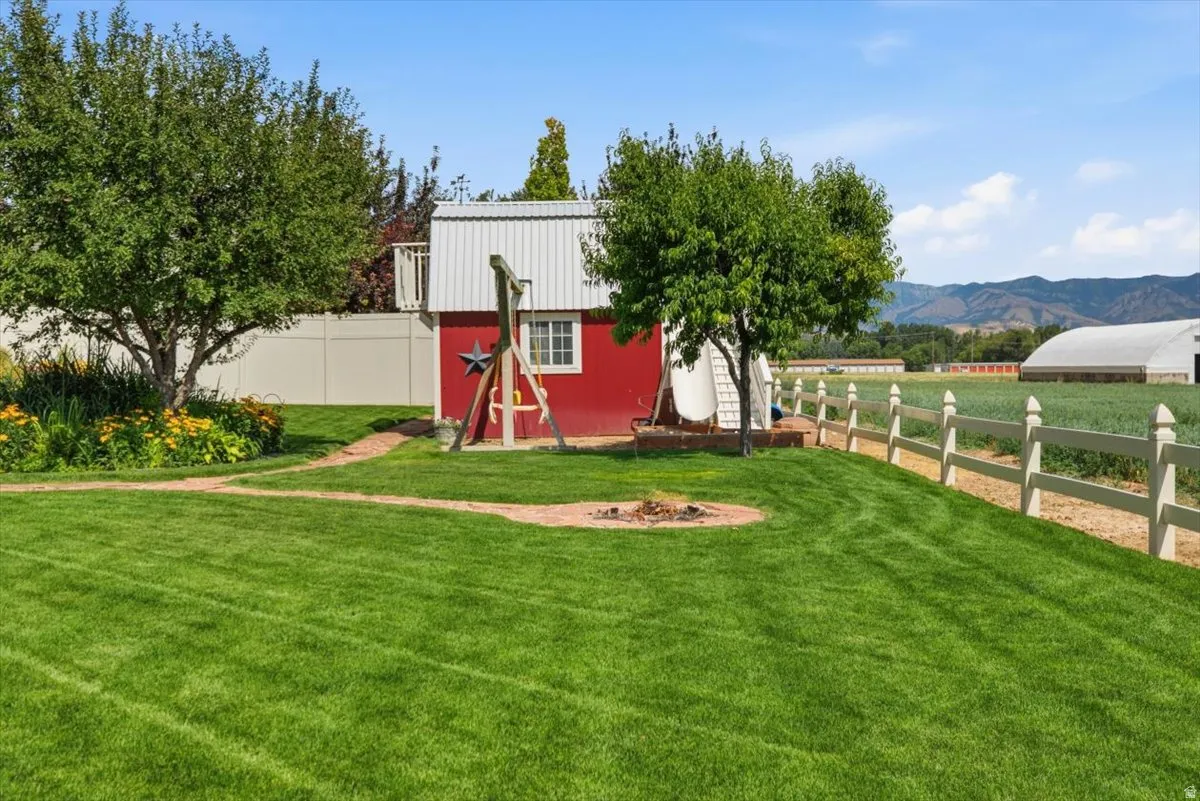 Expansive lawn area featuring a red utility building with white trim, a white privacy fence, mature trees, and a fire pit