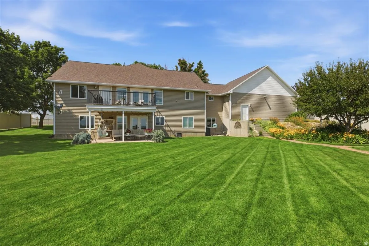 Spacious backyard featuring an upper-level balcony, covered patio, expansive lawn, mature trees, and tiered landscaping with plantings