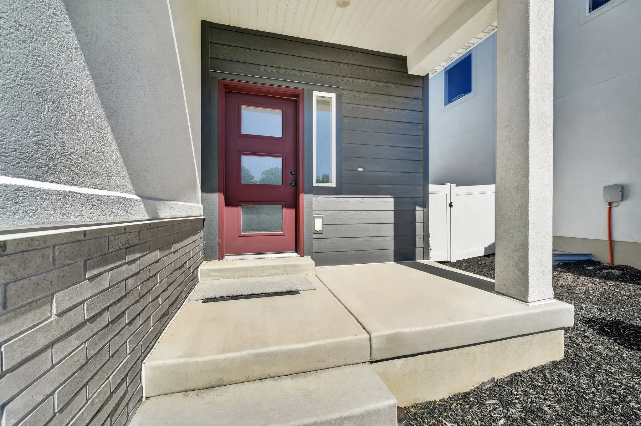 Entryway featuring a dark red door with rectangular glass inserts, concrete steps, a covered porch, dark horizontal siding, and a white fence