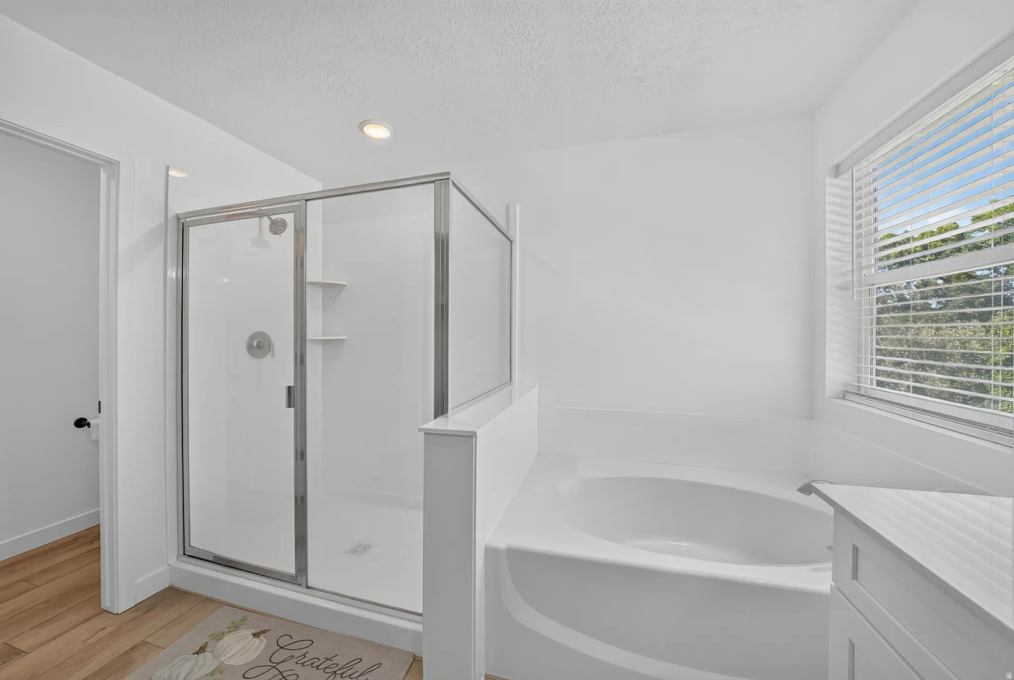 Bathroom featuring a frameless glass shower enclosure, a built-in soaking tub, a window with horizontal blinds, recessed lighting, and wood-finish flooring