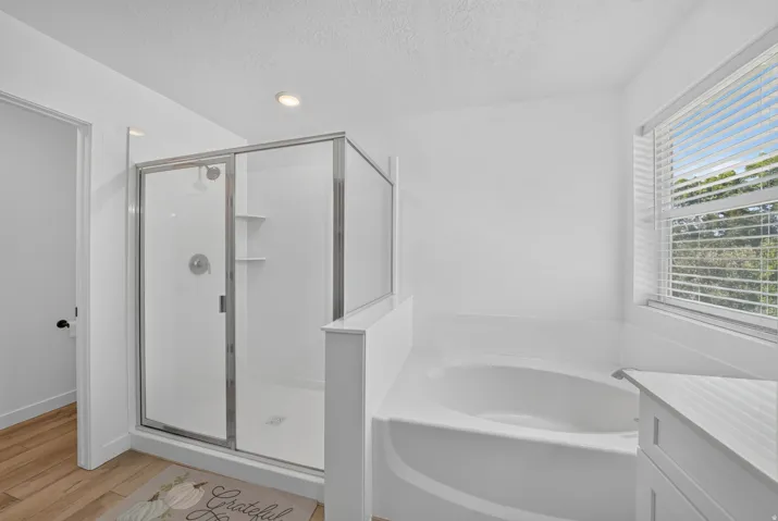 Bathroom featuring a frameless glass shower enclosure, a built-in soaking tub, a window with horizontal blinds, recessed lighting, and wood-finish flooring