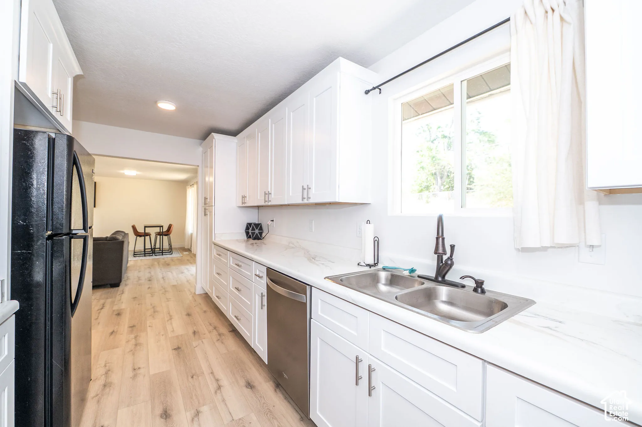 Bright kitchen featuring white shaker cabinetry, stainless steel double-basin sink, bronze-finish faucet, white marble-look countertops, and wood-finish flooring