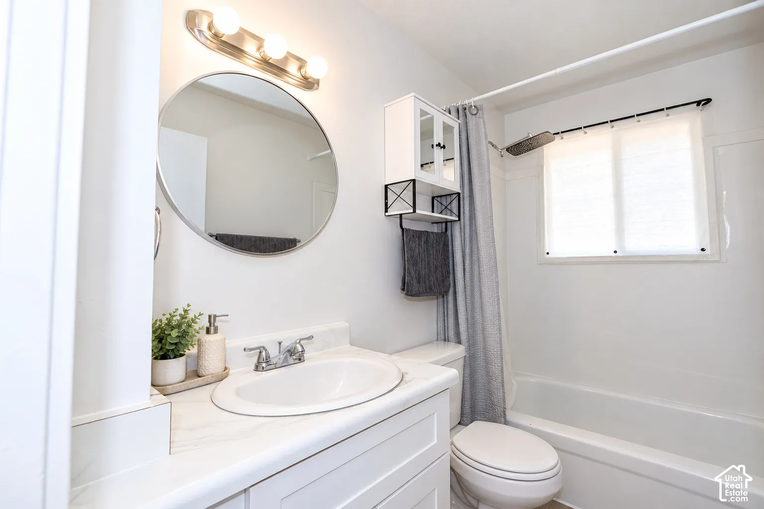 White vanity with an integrated countertop, circular wall-mounted mirror, three-light vanity fixture, and a bathtub with a shower enclosure